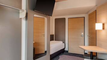 A compact hotel room with a bed, a small desk, a TV mounted on the wall, and a bathroom door.