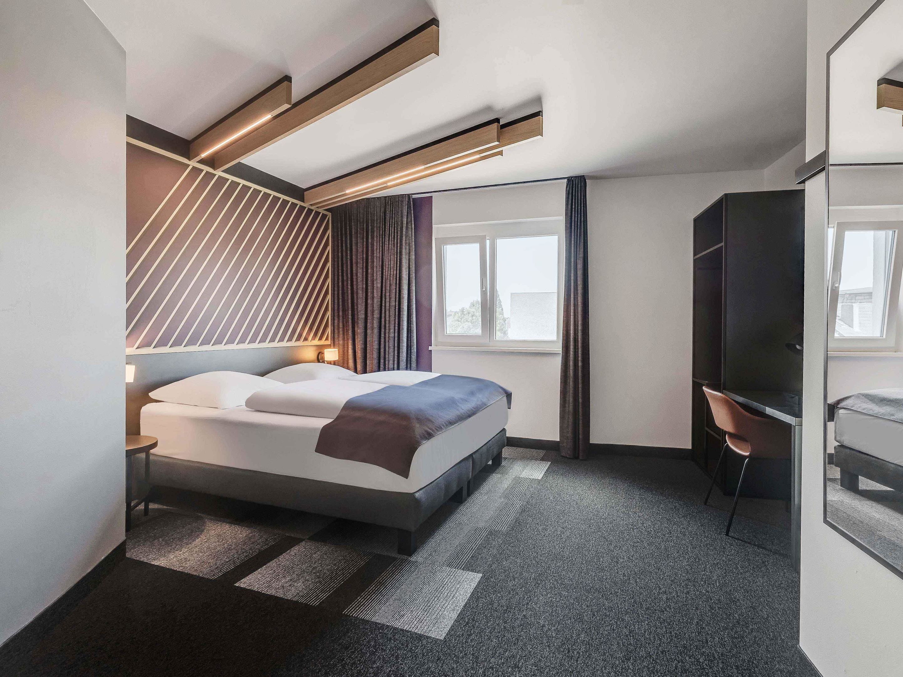 Une chambre d’hôtel moderne, équipée d’un lit, d’un bureau et d’une grande fenêtre.