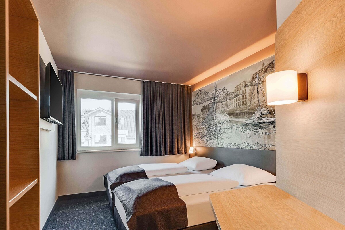 A hotel room with a bed, a desk, a window with curtains, a wall-mounted lamp, and a painting.