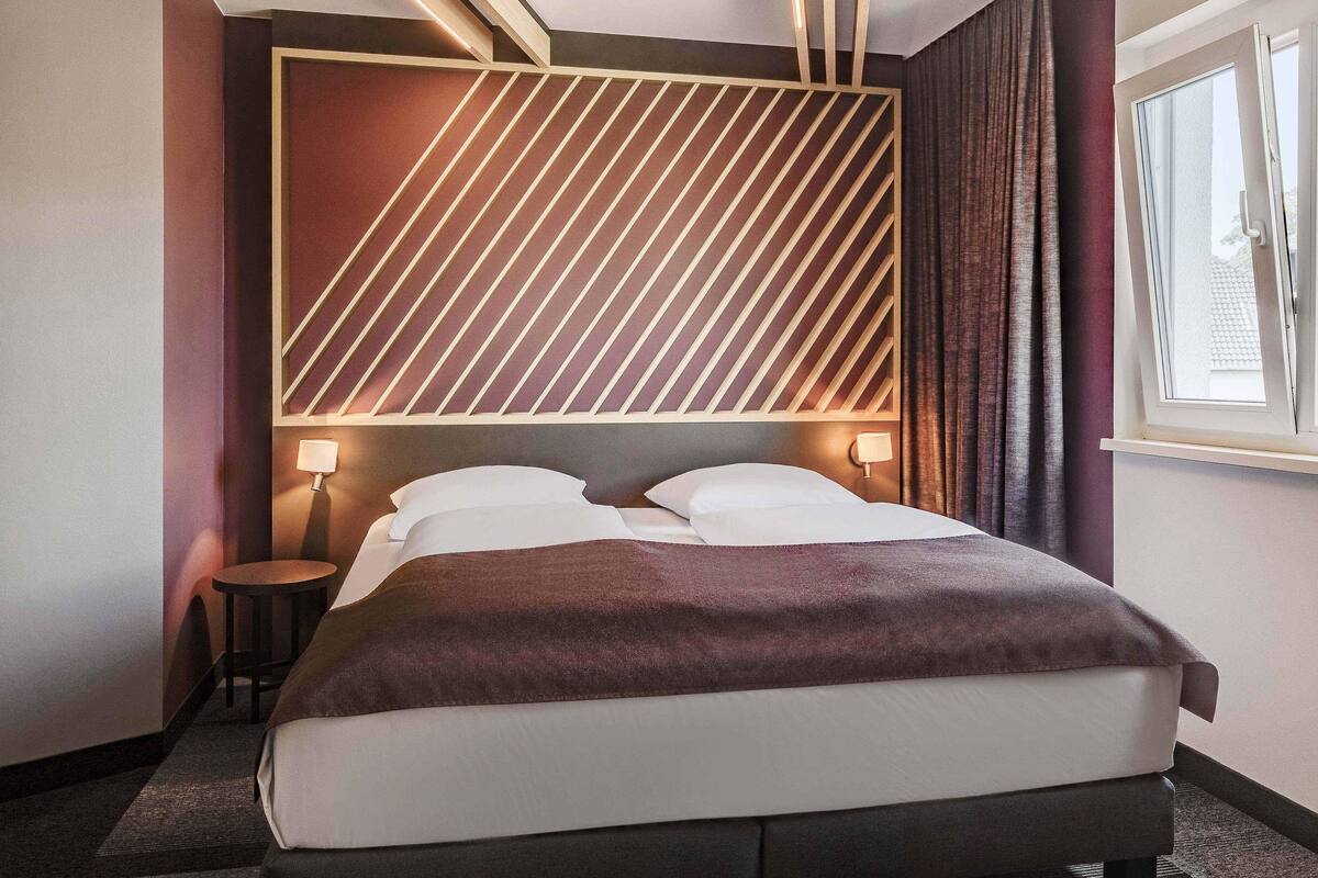 A modern hotel room with a large bed, a headboard with diagonal lines, bedside lamps, and a window with curtains.