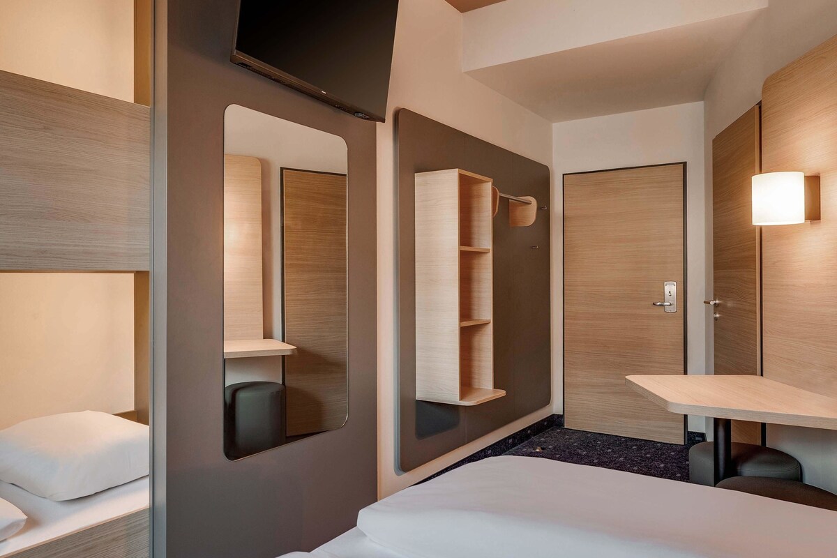 A modern hotel room with a bed, a large mirror, a bedside table, and a wardrobe.