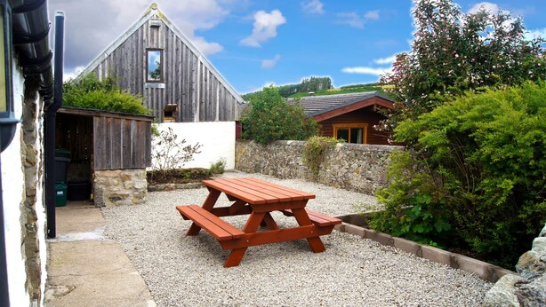 A rustic outdoor area with a wooden picnic table surrounded by gravel, a stone wall, and a wooden building with a sloped roof. - Sunflower Cottage A Cosy, Pet Friendly place to stay. (Huntly)