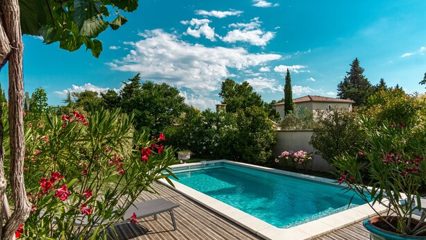 A swimming pool with a wooden deck, surrounded by lush greenery and flowering plants.