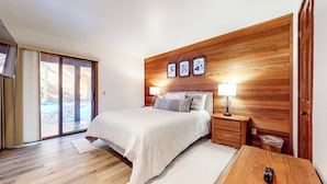 Une chambre à coucher avec une tête de lit en bois, un lit recouvert de linge de lit blanc, deux lampes de chevet, une table de chevet en bois et une porte-fenêtre coulissante donnant sur un espace extérieur.