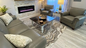 A modern living room with a stone fireplace, leather sofas, a flat-screen TV, and a large window with curtains.