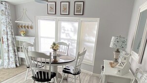 A dining area with a round table, white chairs, a hanging light fixture, and a window with blinds.