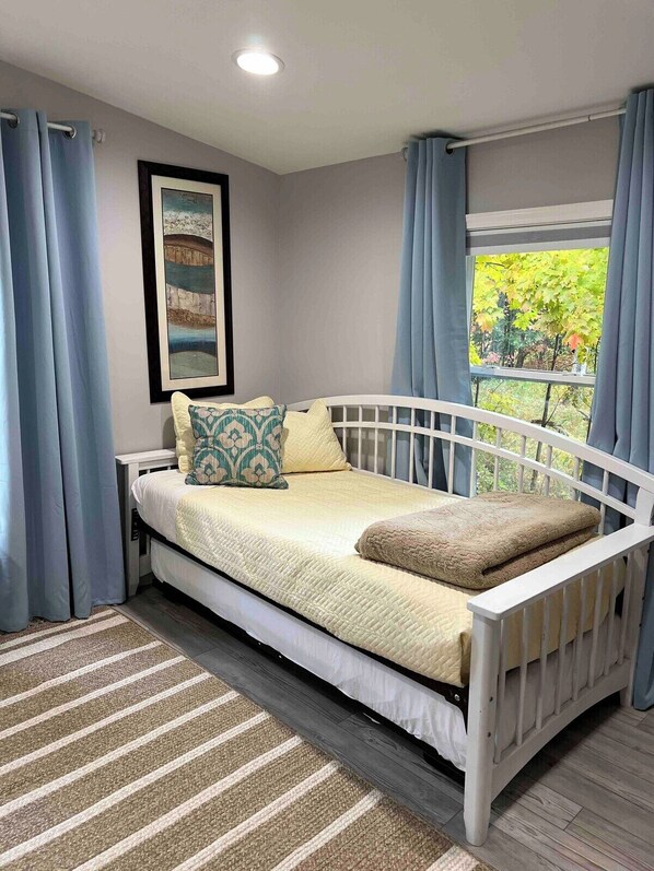 A single bed with a white frame, a beige quilt, and two pillows, a striped rug, a framed picture on the wall, and a window with curtains.