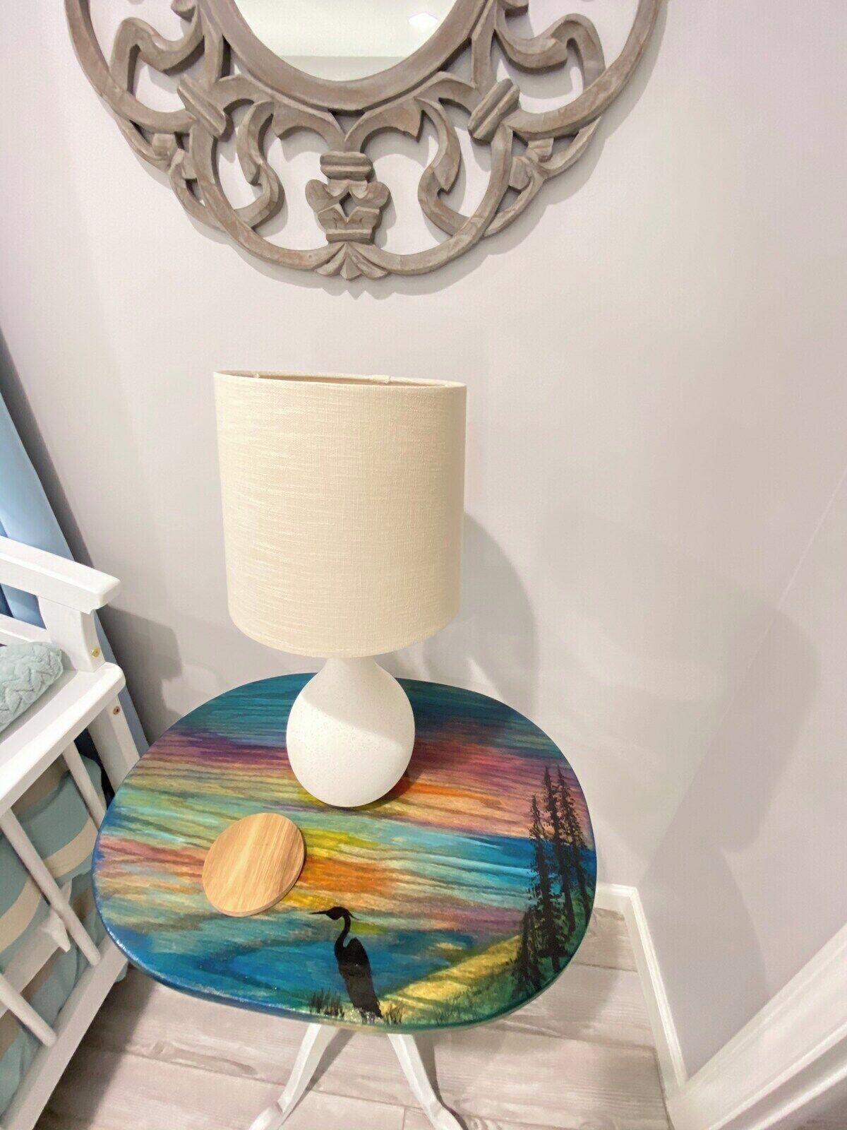 A room with a white lamp on a colorful table, a white crib, and a decorative wall piece.