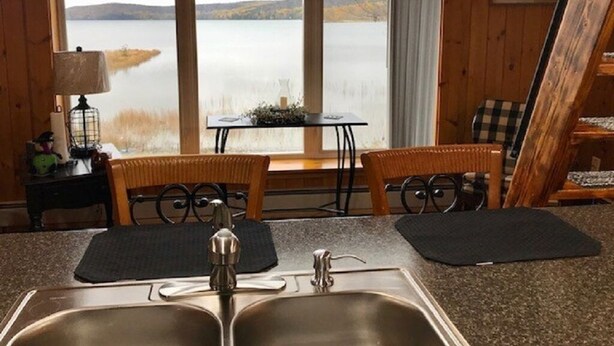 A kitchen with a view of a lake, wooden walls, a sink, and a dining area with chairs and a table. - Direct Trail Access-L & R Lakeside Rentals with this Beautiful Camp on Long Lake (Sinclair)