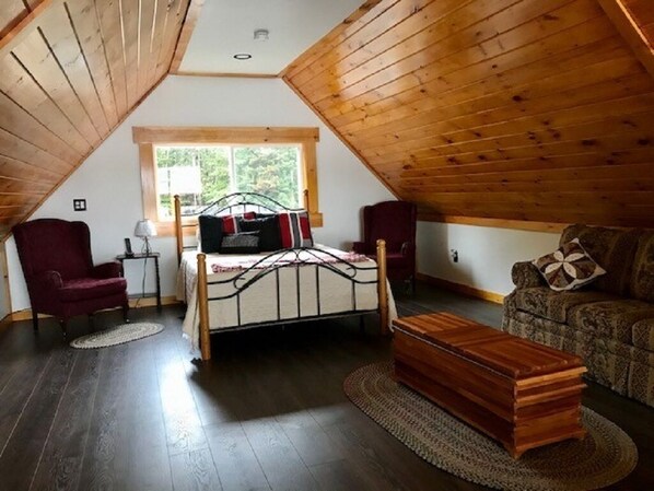 A cozy attic bedroom with a wooden ceiling, a bed, a red armchair, a small table with a lamp, and a patterned sofa. - Direct Trail Access-L & R Lakeside Rentals with this Beautiful Camp on Long Lake (Sinclair)