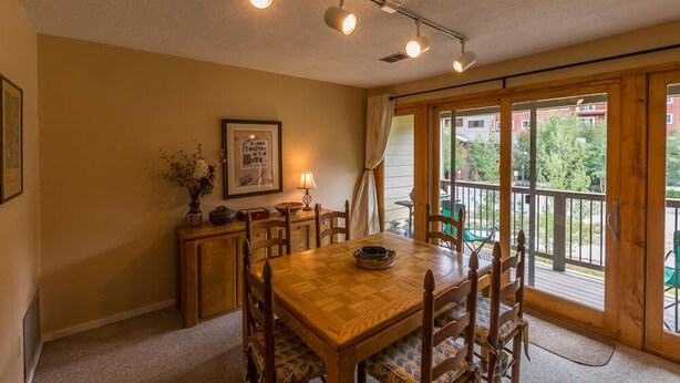 A dining area with a wooden table and chairs, a small cabinet, a framed picture, and a window with a view of greenery. - Alpine Condo | 2 Bedroom | Walk to Lifts | Very Convinient (Mt. Crested Butte)