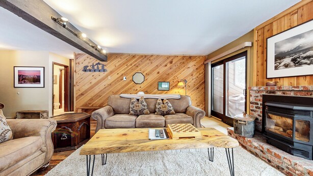 A cozy living room with a wooden wall, a fireplace, a sofa, a coffee table, and a bookshelf. - Spacious Cabin, Rustic Décor. Near Restaurants. Pool & Tennis Court On-site (Mammoth Lakes)