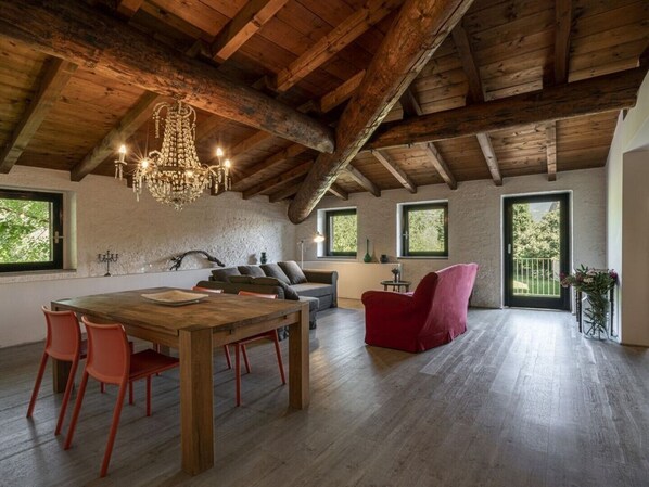 A spacious loft with exposed wooden beams, a wooden dining table with chairs, a red armchair, and a chandelier.