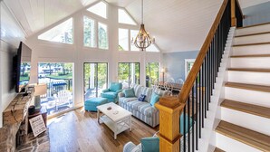 A spacious living room with a high ceiling, large windows, a staircase, and a cozy seating area. - Cozy Cottage at Lake Oconee • Walk-Out Patio • Scenic Porch • Sleeps 8 (Eatonton)