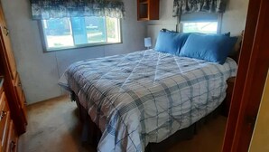 Een slaapkamer met een bed, een plafondventilator, een raam met gordijnen en een klein kastje.