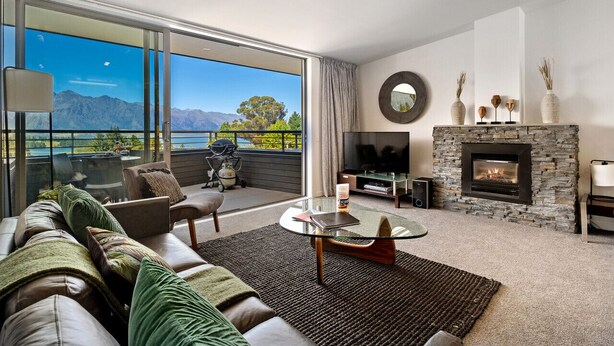 Ein modernes Wohnzimmer mit Ledersofa, Glastisch, Kamin aus Stein und einer großen Schiebetür, die auf einen Balkon mit Blick auf Berge und einen See führt.