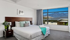 Ein modernes Hotelzimmer mit einem großen Bett, einem Nachttisch mit Blumen in einer Vase, Blick auf Berge durch ein großes Fenster und einem gerahmten Bild an der Wand.