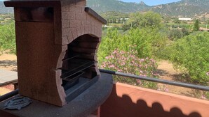 A stone-built outdoor grill with a metal rack, set against a backdrop of green trees and mountains. - Casa Marina (Sardegna)