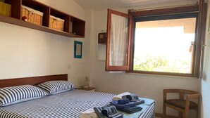 A bedroom with a bed, a chair, a window with curtains, and shelves with baskets. - Casa Marina (Sardegna)
