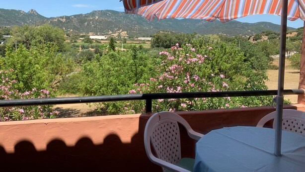 A balcony with a table and chairs, an umbrella, and a view of green hills and pink flowers. - Casa Marina (Sardegna)