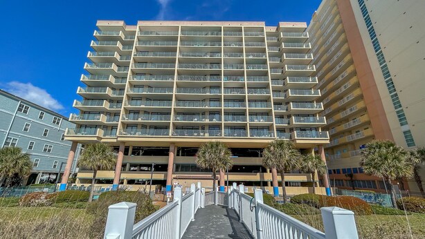 A multi-story hotel with a central entrance, a covered parking area, and a white fence leading to the entrance. - 20% off weekly stays thru Sept. Sat to Sat. 4BR OF condo. Ocean Drive.  Sleeps 12. (North Myrtle Beach)