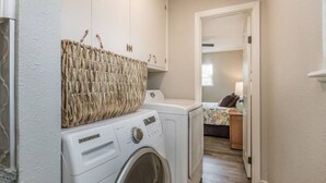 Un espace buanderie équipé d’un lave-linge et d’un sèche-linge, un panier en osier accroché au mur, et une chambre à coucher visible à travers une porte ouverte.
