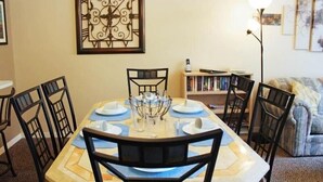 A dining area with a table set for six, surrounded by chairs, a decorative wall piece, and a bookshelf in the background. - Condo Disney (Kissimmee)