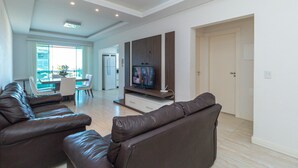 A modern living room with a brown leather sofa, a flat-screen TV, and a dining area with a table and chairs. - Zancanaro 102- Apto - 641B (Bombinhas)