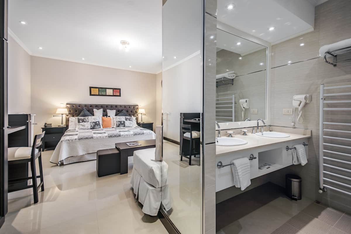 Ein modernes Hotelzimmer mit einem großen Bett, Nachttischen, einem Schreibtisch, einem Stuhl, einem Schminktisch mit Spiegel und einem Badezimmer mit Waschbecken, Handtuchhalter und einer Dusche.