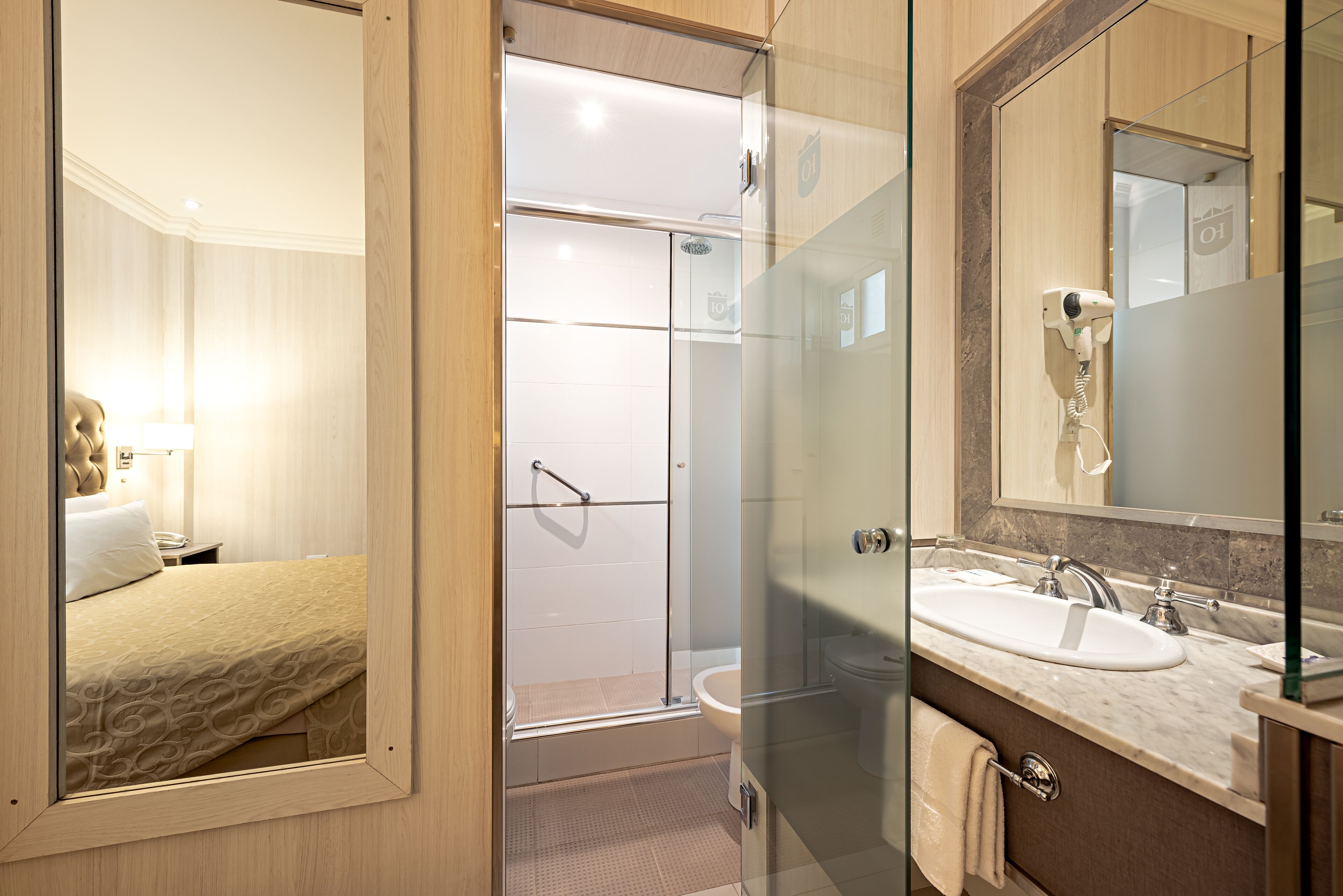 A hotel bathroom with a glass shower enclosure, a sink with a mirror, and a bed with a headboard.