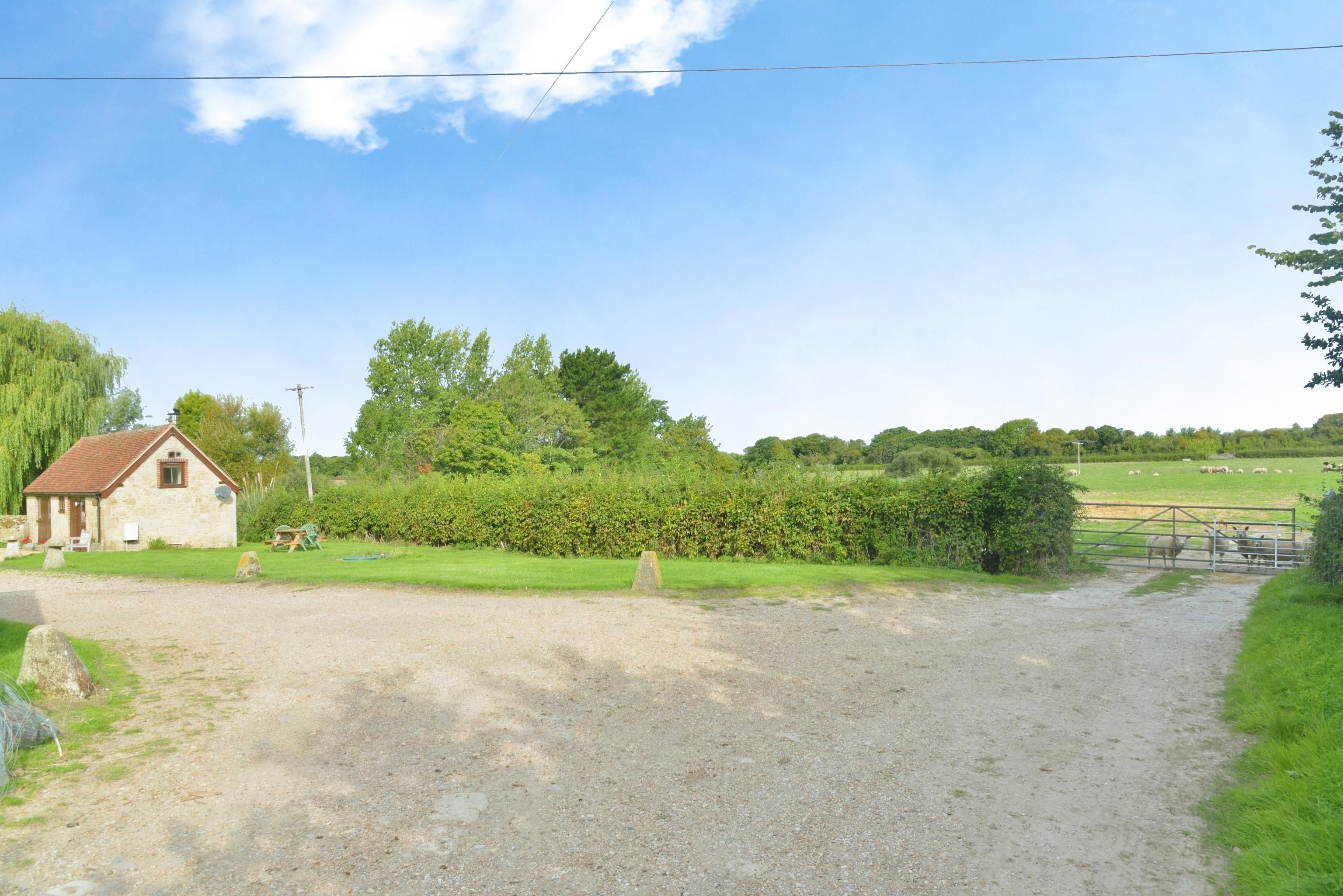 A rural setting with a gravel path leading to a small stone building, surrounded by greenery and a fenced pasture.