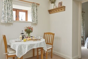 Spisebord med ternet dug, træstole, en vase med blomster, en kande og morgenmadsprodukter.