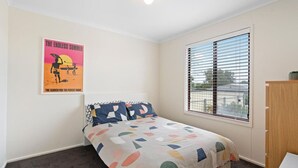 Une chambre à coucher avec un lit, une fenêtre avec des stores, une affiche accrochée au mur et une petite armoire en bois.