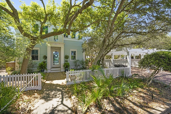 A light green house with a white picket fence, surrounded by lush greenery and trees. - Beachside in Seaside, FL! Amazing Gulf Views and Outdoor Spaces + 2 Bikes! (Santa Rosa Beach)