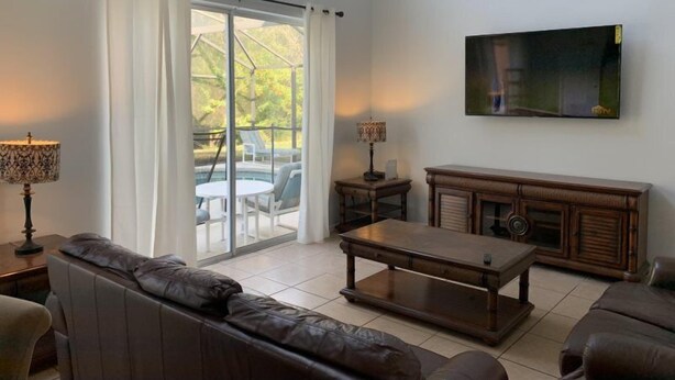 A living room with a sofa, coffee table, TV, and a view through a glass door to an outdoor area with a pool. - Terra Verde 693 (Kissimmee)
