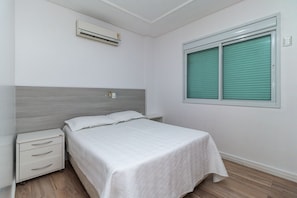A bedroom with a bed, a nightstand, a wall-mounted air conditioner, and a window with blinds. - Zancanaro 203-Apto 641E (Bombinhas)