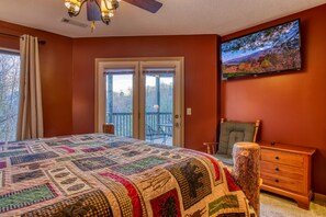 A bedroom with a large bed, a wooden dresser, a television, and a balcony with a view of trees. - Tree Tops by HoneyBearCabins 3/3.5 Sleeps 10 (Gatlinburg)