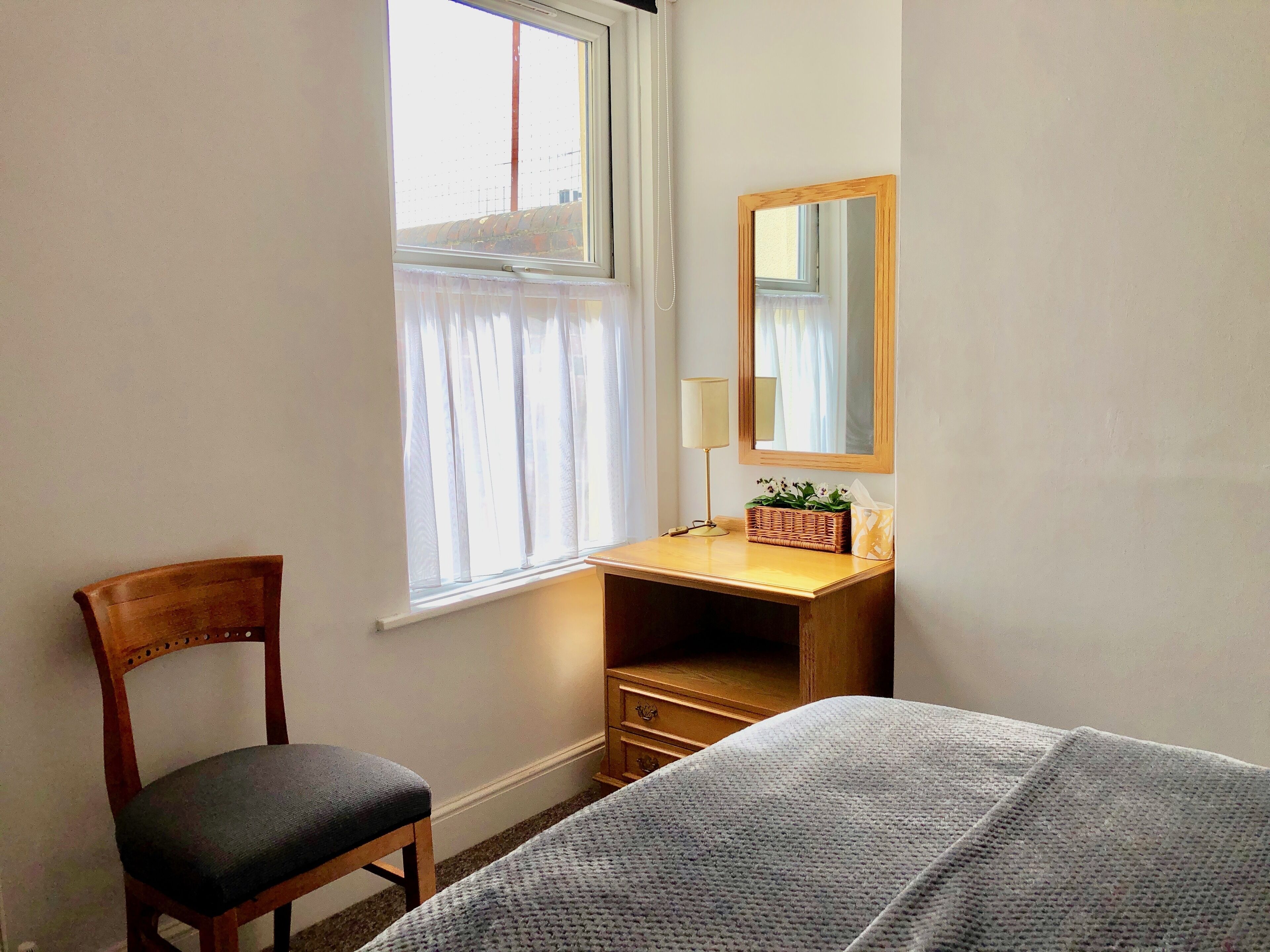 A bedroom with a wooden desk, a chair, a bed with a grey blanket, a bedside lamp, a mirror, and a window with sheer curtains.