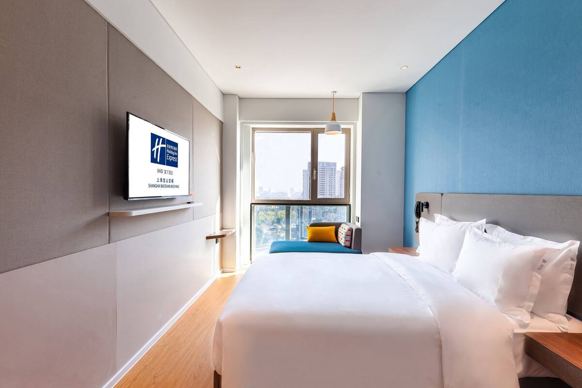 A hotel room with a bed, a TV on the wall, a window with a city view, and a blue accent wall.