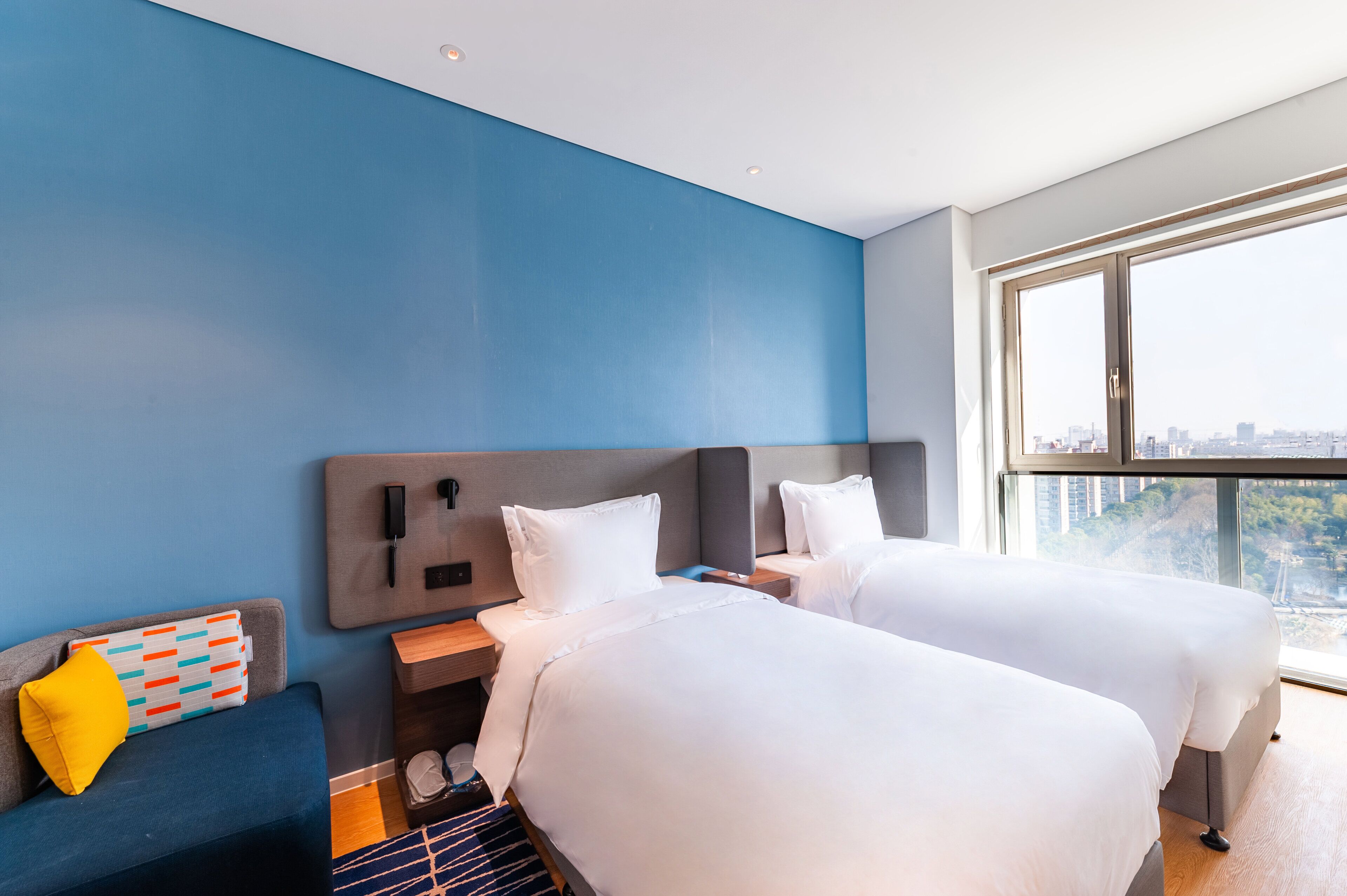 A hotel room with two beds, a desk, a chair, a window with a city view, and a blue accent wall.