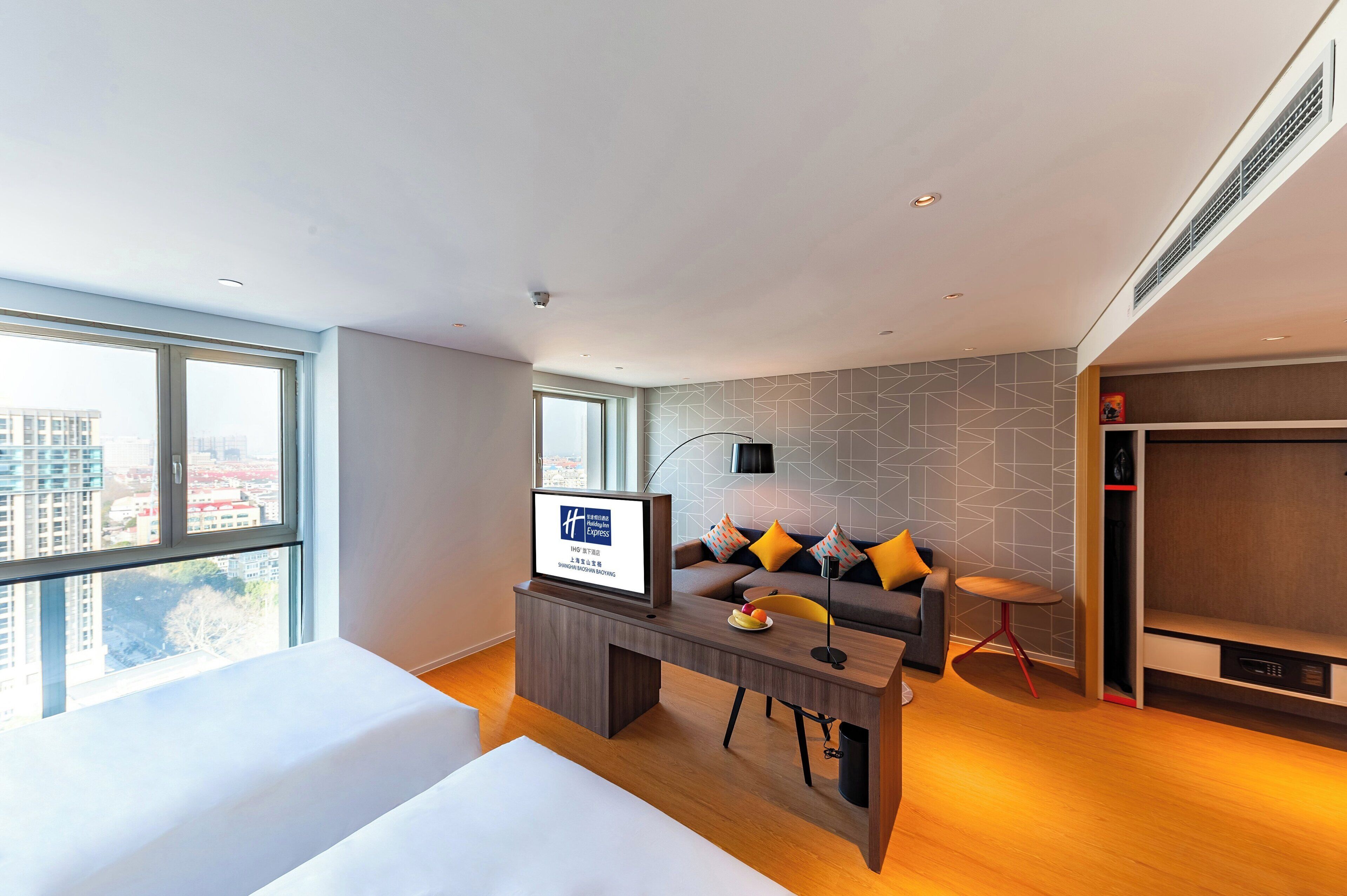 A modern hotel room with a large window, a sofa with cushions, a desk with a laptop, and a television.