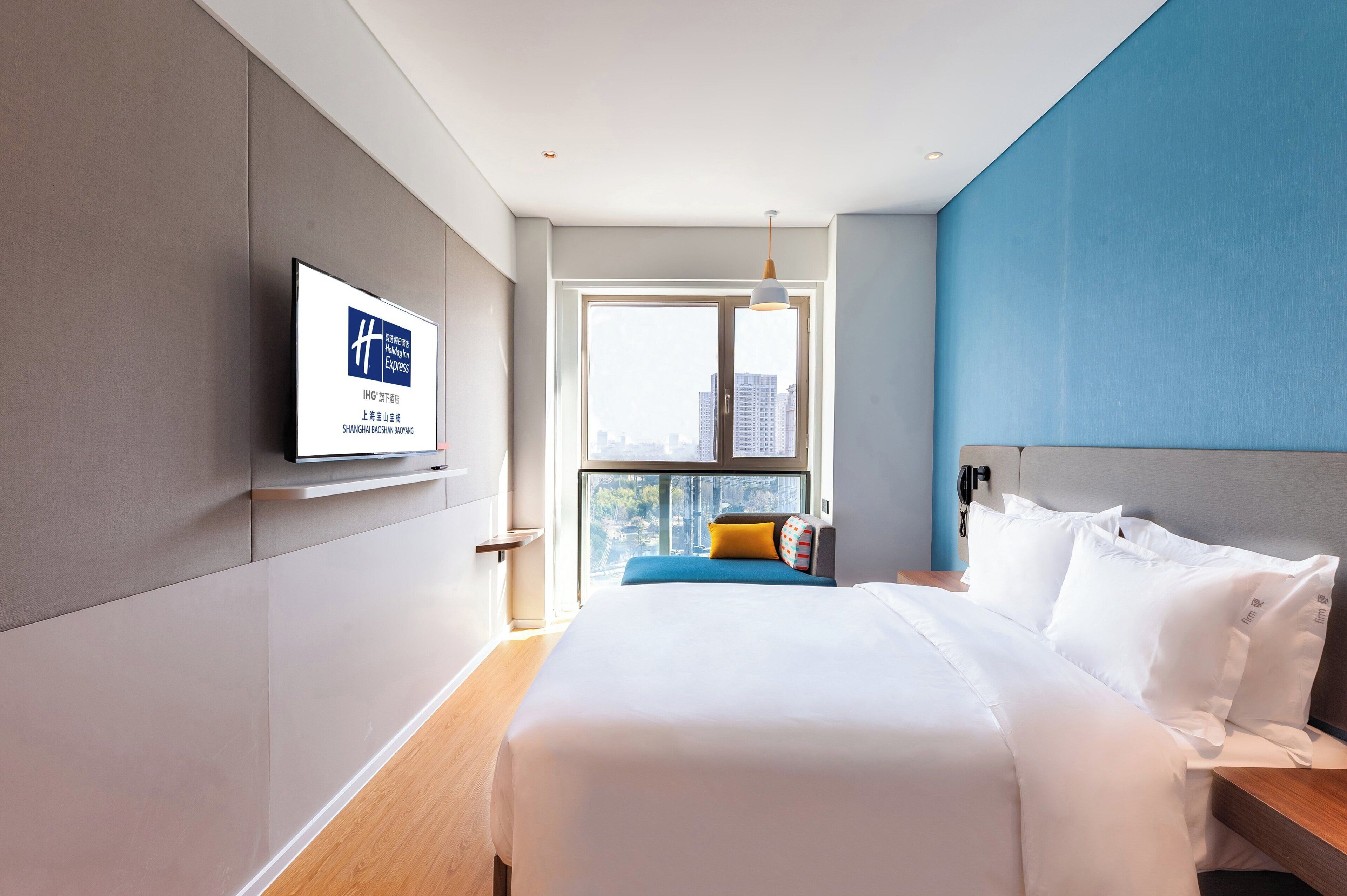 Habitación de hotel con cama, televisor, ventana con vistas a la ciudad y una pared de acento azul.