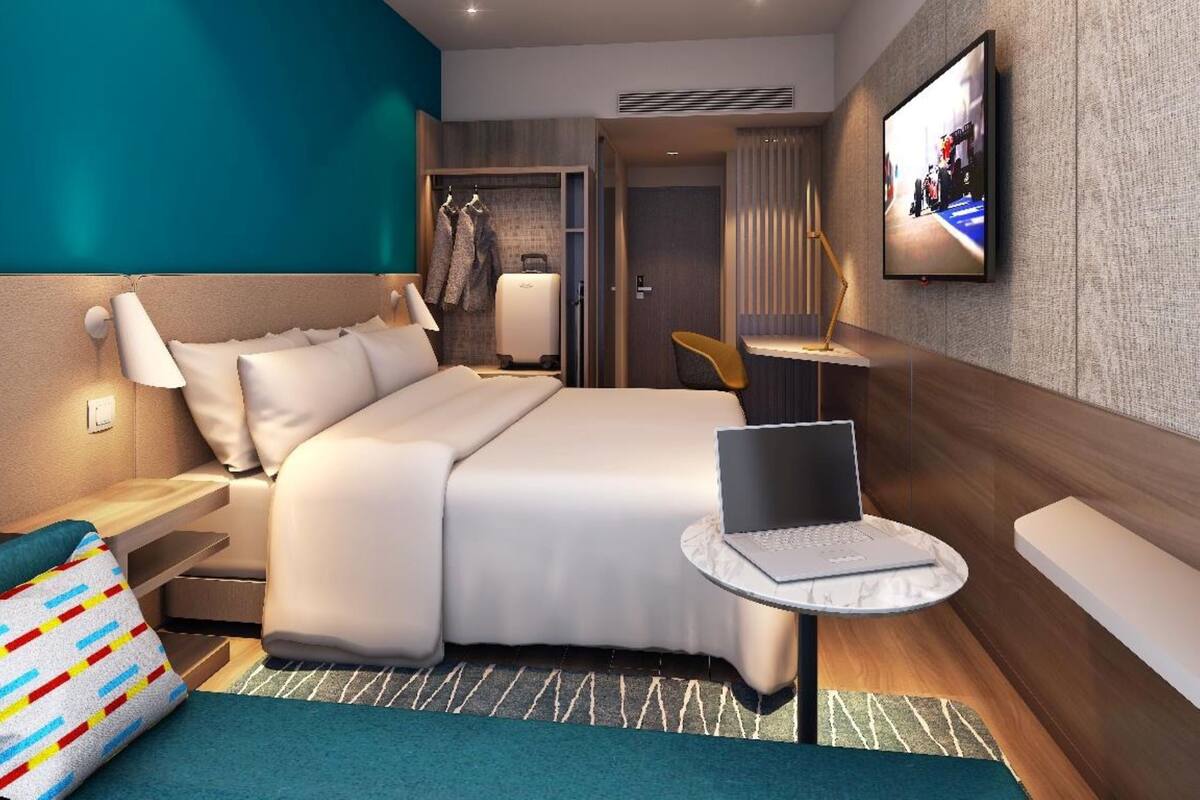 A modern hotel room with a large bed, a desk with a laptop, a TV mounted on the wall, and a wardrobe.