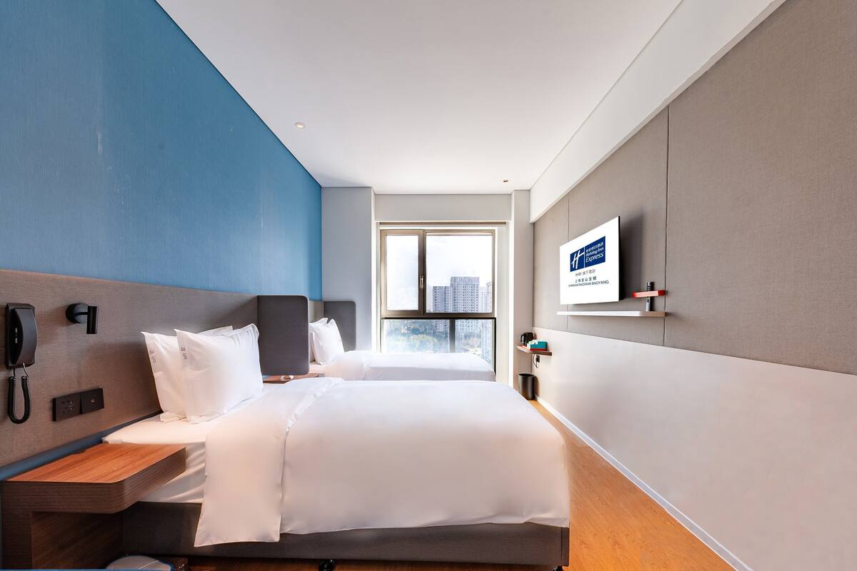 A modern hotel room with a large bed, a bedside telephone, a flat-screen TV mounted on the wall, and a window offering a city view.