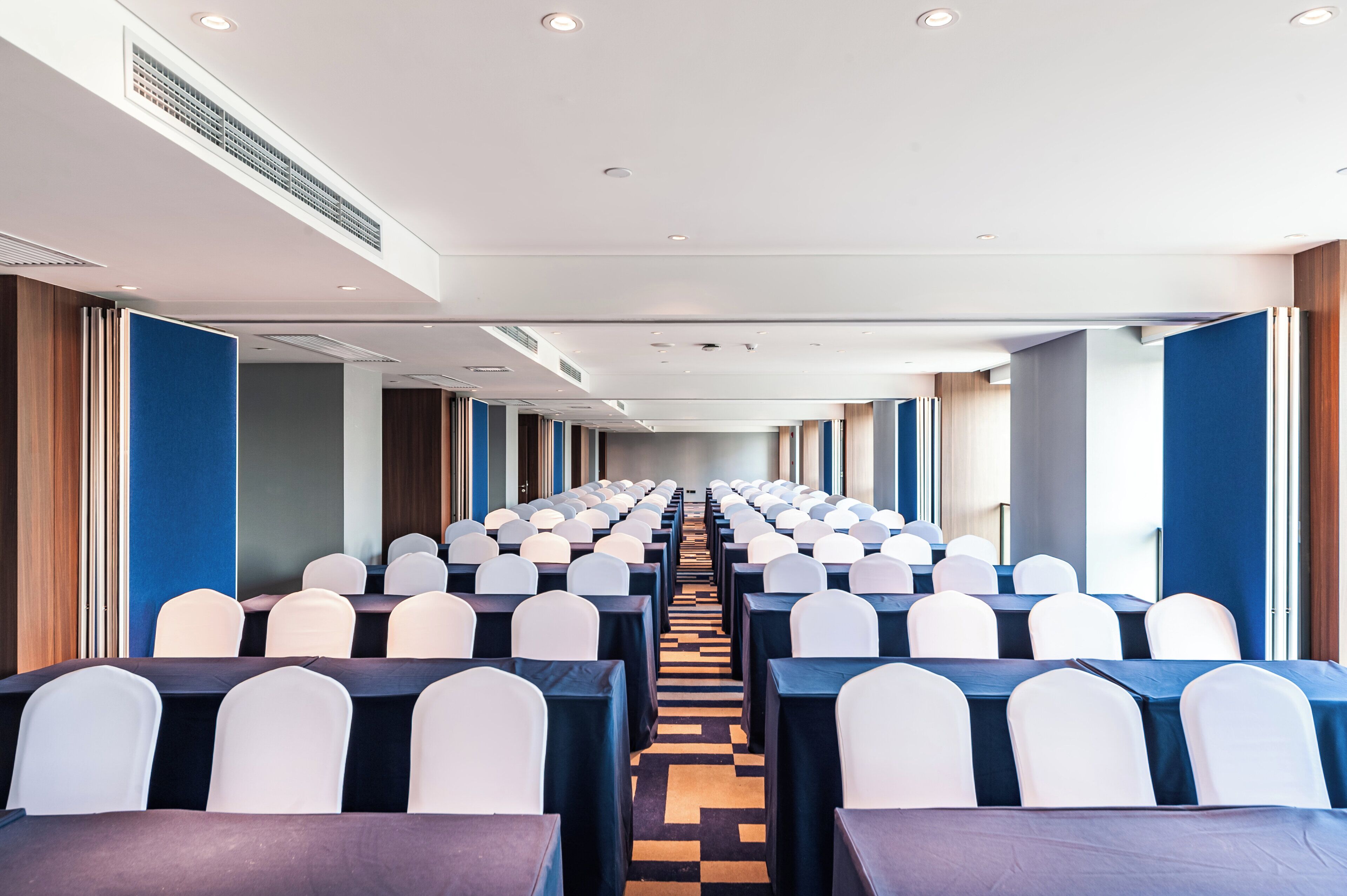 A conference room with rows of tables and chairs, a patterned carpet, and a high ceiling with recessed lighting.