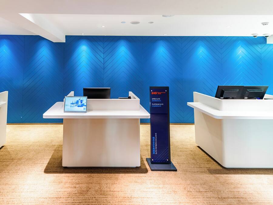 Un área de recepción con tres mostradores blancos, una mesa central con pantalla digital y una pared azul con diseño en forma de chevron.