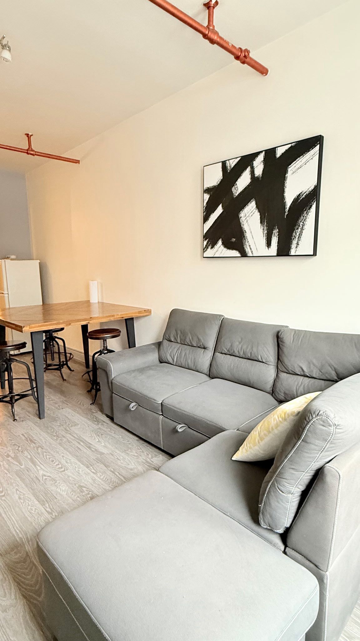 A modern living space with a grey sectional sofa, a wooden dining table with stools, and a black abstract painting on the wall.