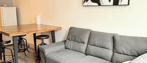 A modern living space with a grey sectional sofa, a wooden dining table with stools, and a black abstract painting on the wall.