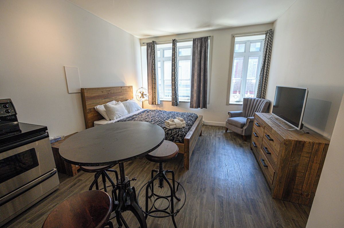 Une chambre à coucher comprenant un lit, une télévision, une table avec des tabourets, une commode et une fenêtre avec des rideaux.