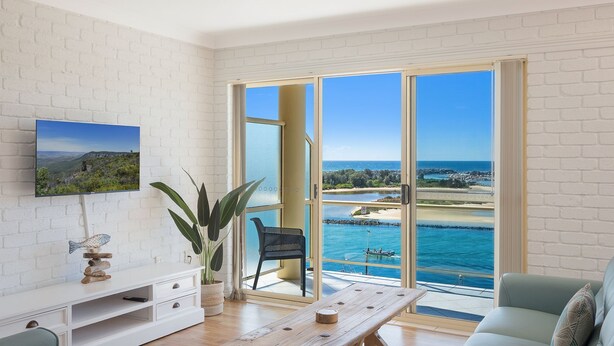 Un salon avec un téléviseur, une plante, une table en bois et une vue sur la mer, visible à travers de grandes portes vitrées.